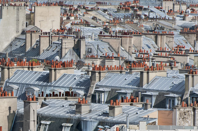 A view looking southwest over buildings in Paris, seen from Basilica Sacré-Cœur: tin rooftops and forests of clay chimney pipes; in the distance is the rooftop of gare du Nord.