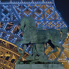 An equestrian statue of a Greek warrior in front of flashing white lights on the Eiffel Tower at night.
