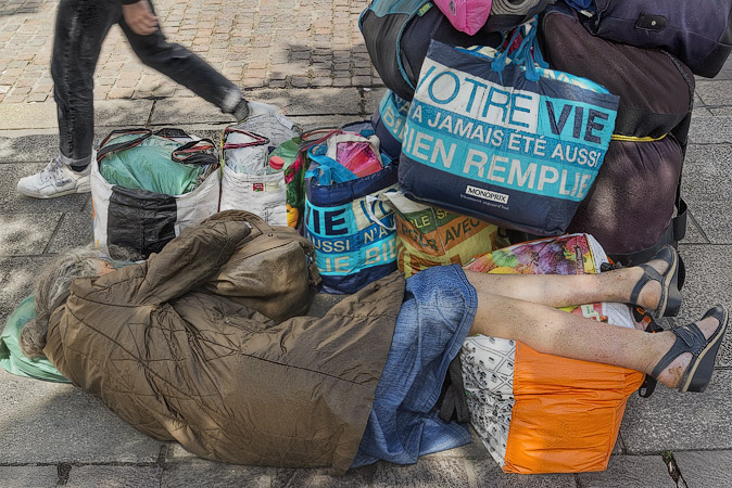 A homeless woman in place Joachim-du-Bellay with Monoprix shopping bags with their slogan, “Your life has never been so full”.
