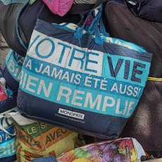 A homeless woman in place Joachim-du-Bellay with Monoprix shopping bags with their slogan, “Your life has never been so full”.
