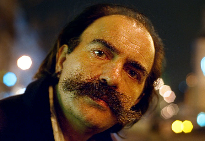 A homeless man with a huge moustache next to place de la Bastille at night.