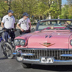 Two policemen on bicycles telling the driver of a pink Cadillac that he can’t remain parked on the Champs-de-Mars.